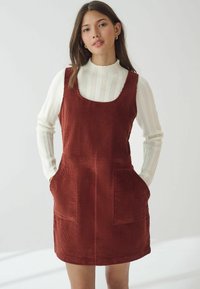 Rustfarbene Cord-Overall-Kleid mit quadratischem Ausschnitt, zwei Fronttaschen und geradem Saum, kombiniert mit einem gestrickten weißen Langarmoberteil.