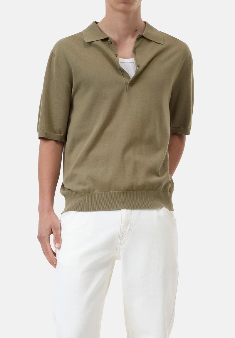 Polo shirt verde oliva a maniche corte con colletto, due bottoni e orlo a costine, abbinato a pantaloni bianchi. Tessuto morbido, vestibilità rilassata.