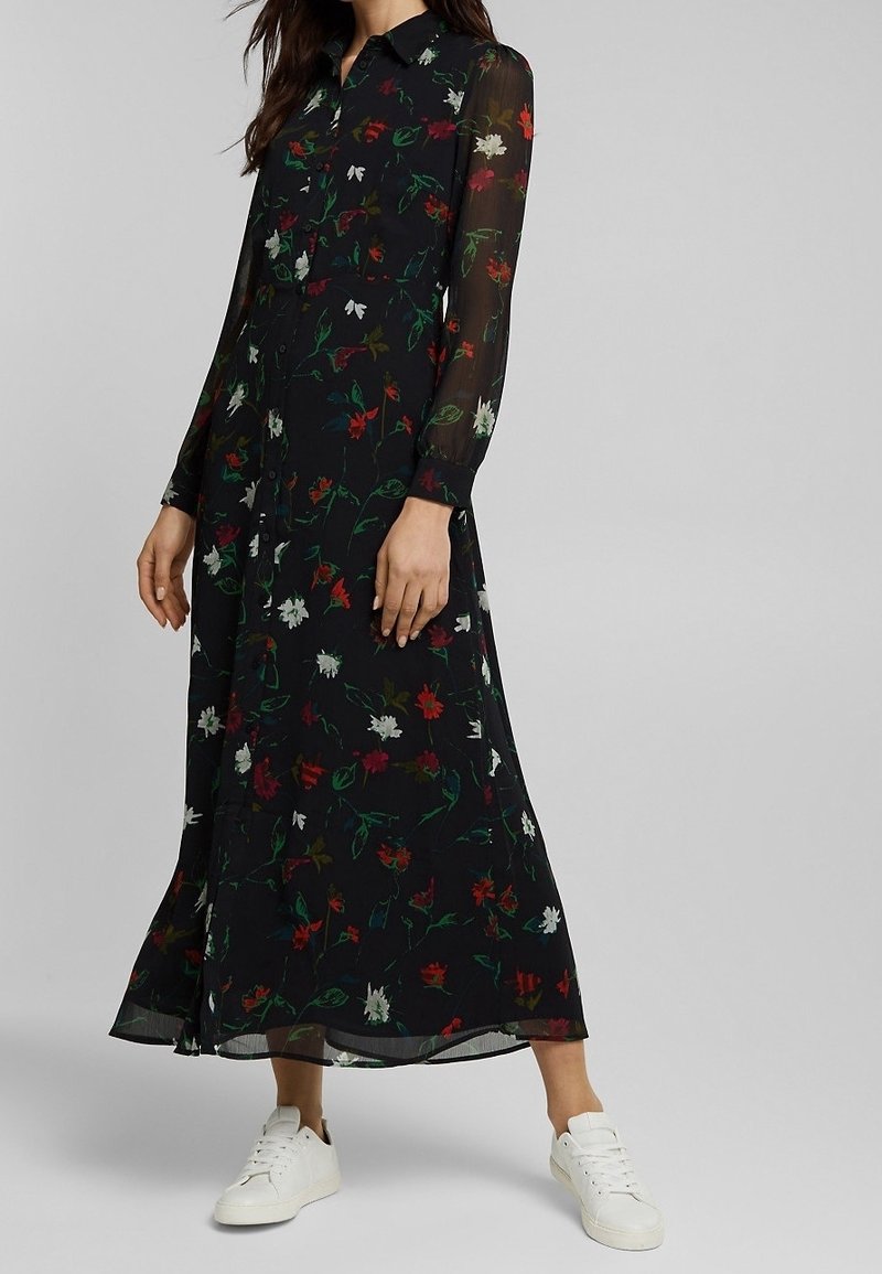 Femme portant une longue robe noire à fleurs avec des manches transparentes et des baskets blanches, debout devant un fond gris uni.