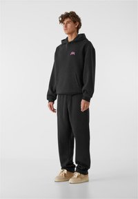 Felpa nera con logo rosa, tasca frontale e pantaloni della tuta abbinati. I materiali sembrano morbidi; le sneaker sono beige con accenti crema.
