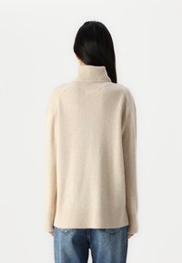 Pull à col roulé beige avec une texture, des poignets et un ourlet côtelés, et une coupe décontractée. Assorti à un jean en denim bleu.