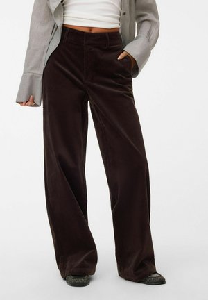 Personne portant un pantalon en velours côtelé marron foncé taille haute à jambes larges, un top court blanc, une chemise grise oversize et des mocassins noirs, main dans la poche.