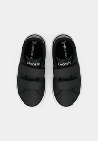 Sneakers neri con due cinturini in Velcro, tomaia in sintetico liscio, suole in gomma bianca e logo Lacoste sulla linguetta e sul lato.