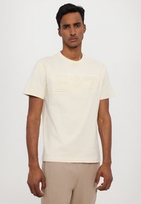 Beige katoenen T-shirt met een verhoogd "EA7" logo aan de voorkant, korte mouwen en een ronde halslijn, gecombineerd met lichtgekleurde broek.