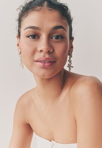 Ohrringe mit floralem Design aus goldfarbenem Metall und filigranen Details. Modell hat dunkle Haare und einen natürlichen Make-up-Look.