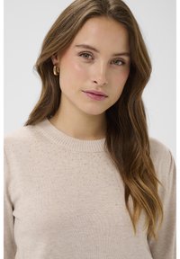 Bež pleten pulover z rebrastim izrezom in pikčastim vzorcem. Model nosi zlate obroče za ušesa, lasje so stilizirani v ohlapne valove.