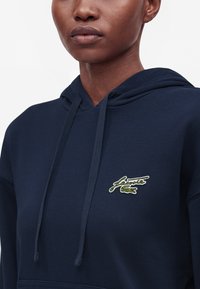 Gros plan d'une personne portant un sweat à capuche bleu marine avec un logo Lacoste vert brodé sur la poitrine.