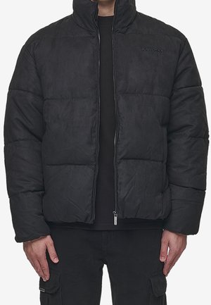 Mand iført en sort polstret lynlåsjakke med høj krave og sorte cargobukser, der står med armene afslappet ned langs siden.