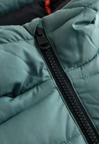 Padded jacket in soft teal fabric with a textured surface. Features a contrasting black zipper, closely stitched seams, and minimal design.