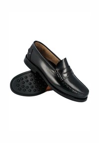 Mocasines de cuero negro con un acabado liso, que presentan una correa decorativa en la parte superior y una suela de goma texturizada con patrones circulares.