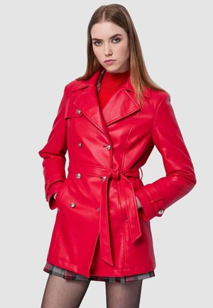 Trench coat doppiopetto in pelle rossa con vita con cintura, accessori in argento e rever pronunciati, indossato sopra un maglione rosso.