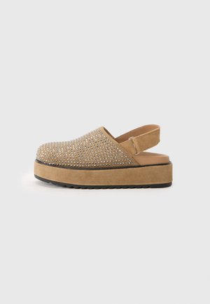 Beige suedeklippere med en teksturert overdel dekket av sølvnagler, åpen bak, og en tykk platåsåle med en svart gummibase.