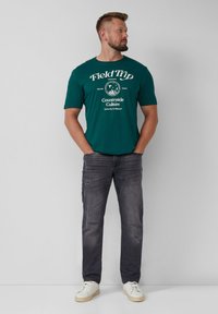 Teal katoenen t-shirt met witte grafische tekst; gecombineerd met donkere, licht versleten jeans en beige sneakers. Eenvoudige, casual outfit.