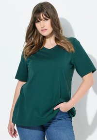 Zelené tričko s výstřihem do V vyrobené z měkké látky, s krátkým rukávem a volným střihem, kombinované s modrými džíny, na světlém pozadí.
