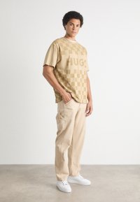 Camiseta de cuadros beige con el texto "HUGO", combinada con pantalones cargo de color beige claro y zapatillas blancas. El conjunto tiene un ajuste relajado.