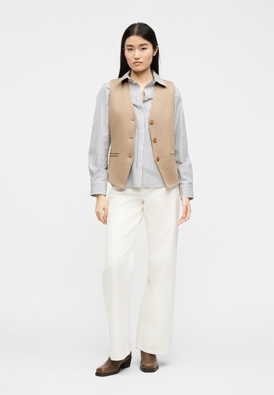 Γυναίκα με μπεζ γιλέκο με κουμπιά πάνω από ριγωτό πουκάμισο, λευκό παντελόνι με φαρδιά πόδια και καστανά μπότες με τακούνι, που στέκεται μπροστά σε απλό φόντο.
