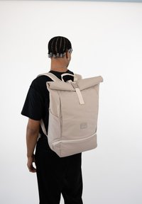 Beige roll-top backpack with adjustable straps, a front zipper pocket, and a logo patch on the lower section. Smooth texture, minimal design.