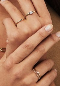 Zwei goldene Ringe an Fingern. Einer hat einen zentralen klaren Stein; der andere ist mit kleinen Perlenakzenten strukturiert. Glatte, polierte Oberfläche.