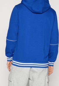 Felpa blu con cappuccio regolabile, polsini e orlo a costine con strisce bianche. Il tessuto sembra morbido e strutturato. Abbinata a pantaloni grigi.
