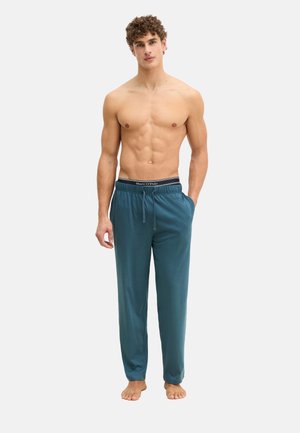 Zonder shirt staande man met krullend haar, blootsvoets, gekleed in teal kleurige broek met trekkoord en zwarte Marc O'Polo onderbroek met tailleband, tegen een witte achtergrond.