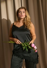 Γυναίκα με μακριά ξανθά μαλλιά που φοράει μαύρο μπλουζάκι χωρίς μανίκια με πουά και παντελόνι, κρατώντας ροζ λουλούδια μπροστά από φόντο μπεζ κουρτίνας.