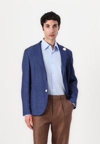 Blue textured blazer with a notched lapel, light blue button-up shirt, and brown pants. Small white flower detail on the blazer's lapel.