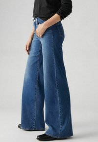 Jeansy o szerokich nogawkach w średnim niebieskim denimu, z wysokim stanem, subtelnym wyblaknięciem i czystym wykończeniem dołu. W połączeniu z czarnymi butami, główny akcent pada na jeansy.