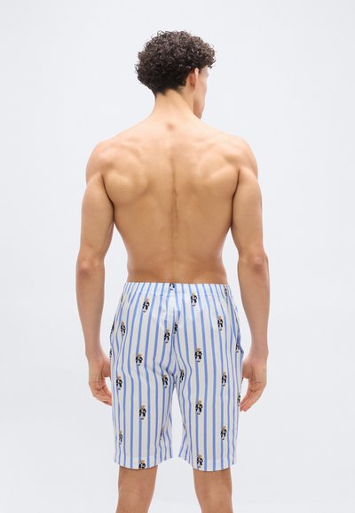 Άνδρας με σγουρά μαλλιά, χωρίς πουκάμισο, φοράει άσπρες και μπλε ριγέ σορτς μεσαίου μήκους με μικρά σχέδια αρκούδων, στέκεται μπροστά σε απλό ανοιχτό φόντο.