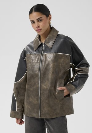 Femme aux cheveux foncés portant une veste en cuir oversize bicolore marron et noir avec un passepoil blanc, debout, une main dans la poche et une expression neutre.
