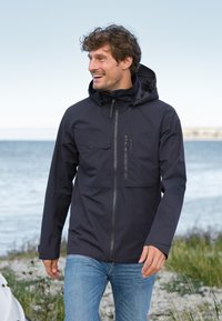 Laivastonsininen vedenpitävä takki, jossa on edessä vetoketju, rintatasku ja säädettävä huppu. Yllä siniset farkut, taustalla rannikkonäkymä.