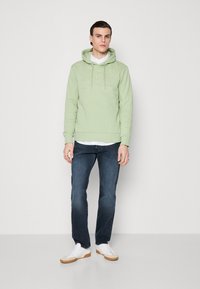 Sudadera verde claro con bolsillo tipo canguro y logotipo en relieve, combinada con jeans de mezclilla oscura y zapatillas blancas con suela de goma.