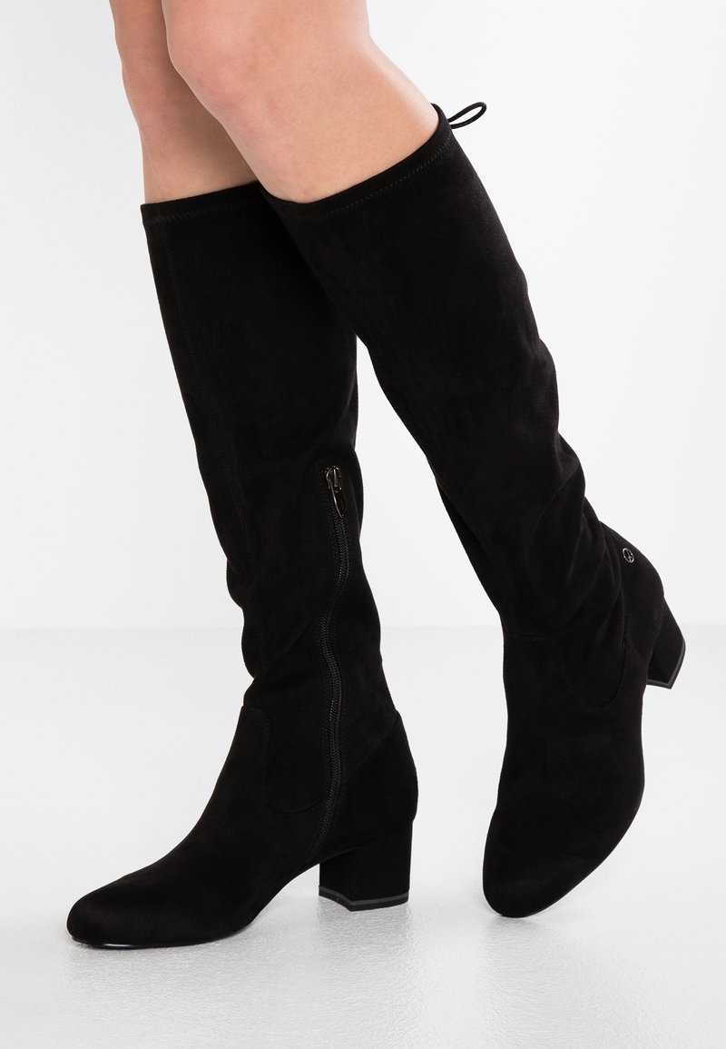 Bottes montantes en daim noir avec talons blocs et fermetures éclair latérales portées sur des jambes croisées sur fond blanc.