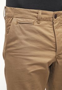 Pantalones caqui con una textura suave, que cuentan con un único bolsillo ribeteado y un cierre de botón en la cintura. Vista en primer plano.