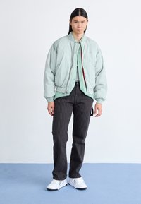 Jaka mint zelene boje, bomber stil, preko svijetle košulje s gumbima, u kombinaciji s crnim hlačama s prugama i bijelim tenisicama. Glatka tekstura, opušteni kroj.