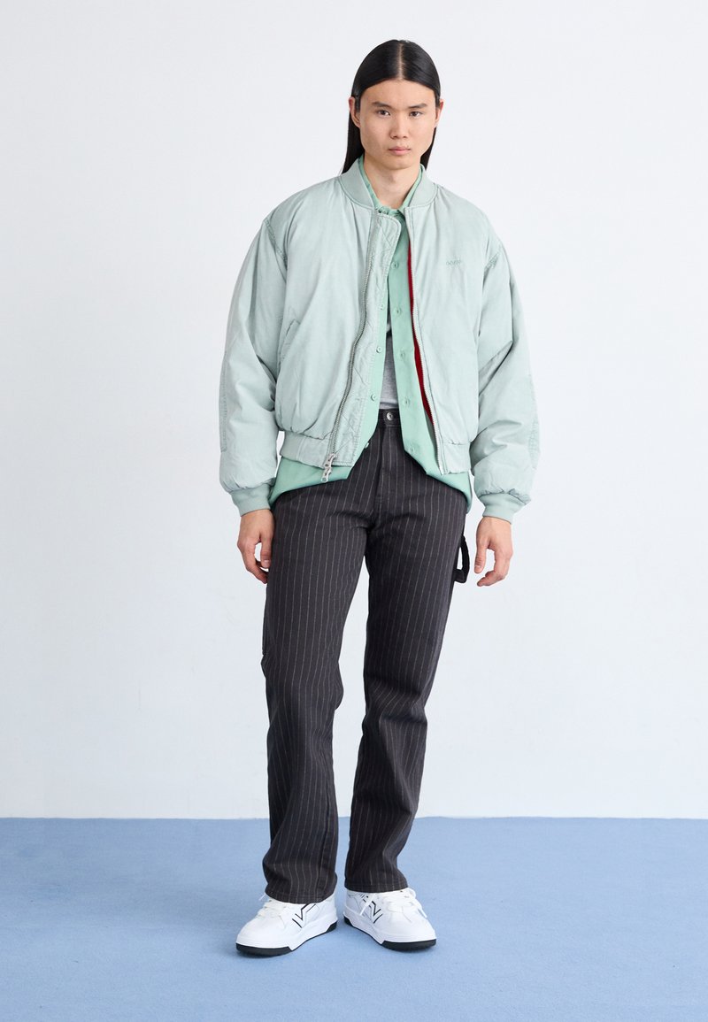 Mintgrüne Bomberjacke über einem hellen Hemd mit Knopfleiste, kombiniert mit schwarzen Nadelstreifenhosen und weißen Sneakers. Glatte Textur, entspannte Passform.