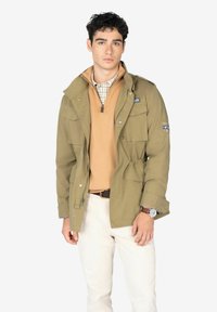 Chaqueta verde oliva con bolsillos frontales y cierre de botones a presión, superpuesta sobre un suéter marrón claro y una camisa a cuadros, combinada con pantalones blancos.