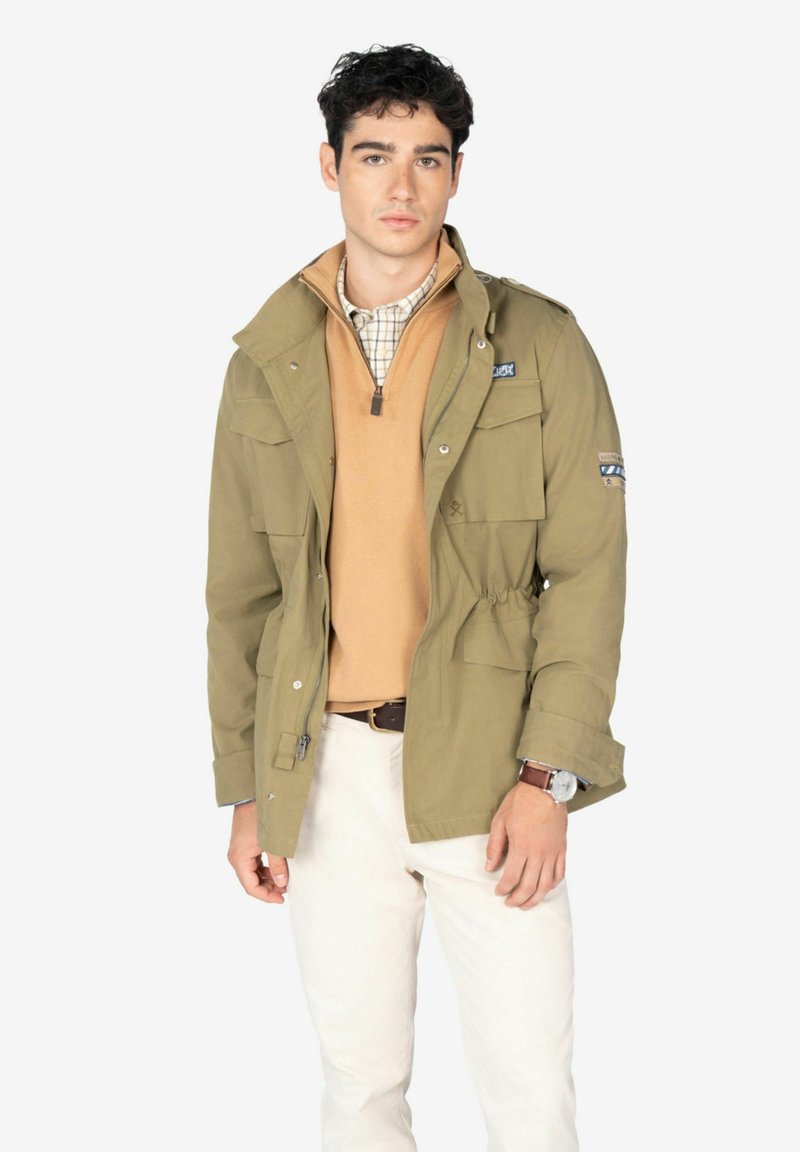 Chaqueta verde oliva con bolsillos frontales y cierre de botones a presión, superpuesta sobre un suéter marrón claro y una camisa a cuadros, combinada con pantalones blancos.