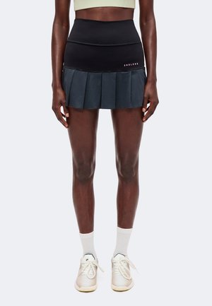 Personne athlétique portant une jupe de tennis plissée noire avec le logo "ENDLESS", un haut de sport clair, des chaussettes blanches et des baskets blanches, debout.