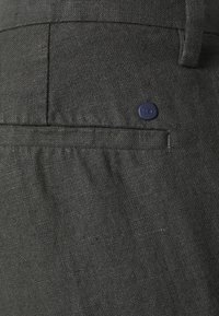 I pantaloni in lino grigio scuro presentano una tasca posteriore con un piccolo bottone blu marina e cuciture visibili. Il tessuto ha una finitura strutturata.