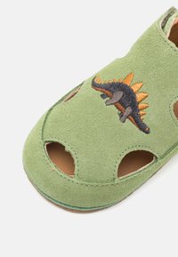 Grüne Wildleder-Sandale mit Ausschnitt-Design und Dinosaurier-Stickerei in Schwarz und Orange oben. Braune Sohle an der Unterseite sichtbar.