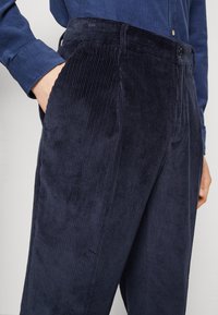 Navy blue corduroy trousers with wide vertical ridges, featuring side pockets and a flat front, accented by a button and zipper closure.