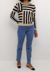 Gebreide pullover in beige en zwart met verschillende gestreepte patronen, gecombineerd met blauwe jeans en twee-kleurige blokhak schoenen.