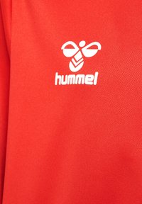 Maglia sportiva rossa con tessuto strutturato. Presenta un logo Hummel bianco con un'ape sopra il nome del marchio in lettere minuscole.