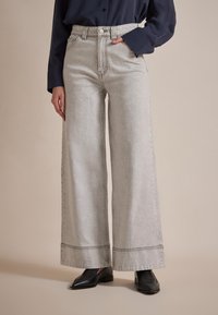 Wijd-uitlopende jeans in lichtgrijs denim, met een hoge taille, rafelige zoom en subtiele stiksels. Gecombineerd met zwarte platte schoenen.