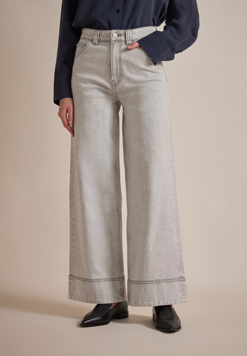 Wijd-uitlopende jeans in lichtgrijs denim, met een hoge taille, rafelige zoom en subtiele stiksels. Gecombineerd met zwarte platte schoenen.