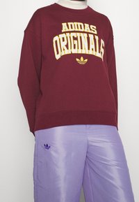 Bordeaux sweatshirt med gul "ADIDAS ORIGINALS" text; rund halsringning och ribbade ärmmuddar, i kombination med glänsande lila byxor med en liten logotyp.