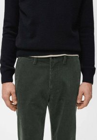 Mężczyzna w ciemnozielonych spodniach sztruksowych i czarnym swetrze z długim rękawem, stojący z rękami swobodnie opuszczonymi wzdłuż ciała na białym tle.