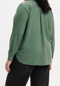 Chemise verte à manches longues en tissu lisse avec une coupe décontractée, un ourlet arrondi et des poignets boutonnés, assortie à un pantalon noir.