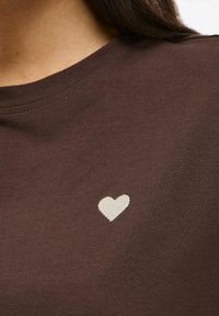 Maglietta in cotone marrone con un piccolo cuore bianco ricamato sulla zona del petto. Tessuto liscio e classico scollo rotondo.