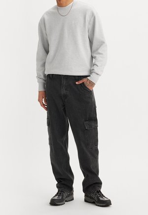 Homme portant un sweat-shirt gris clair, un jean cargo noir, des baskets noires et grises, avec une main tatouée dans la poche, debout sur un fond blanc uni.